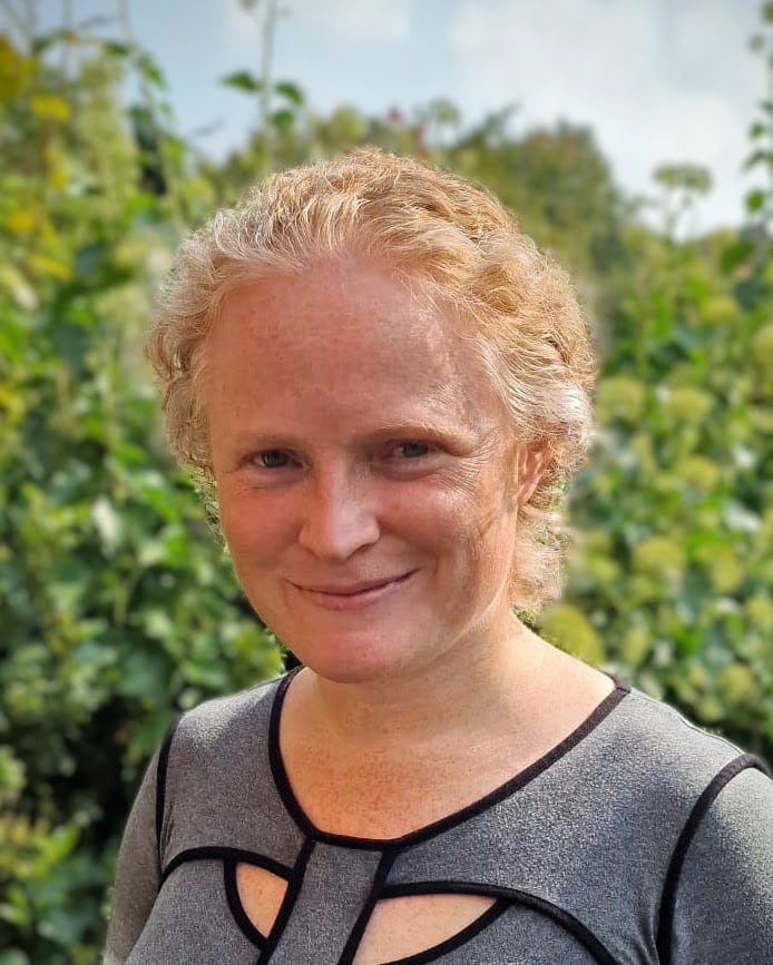 A headshot of Bridget Penman standing outdoors in front of a green leafy background