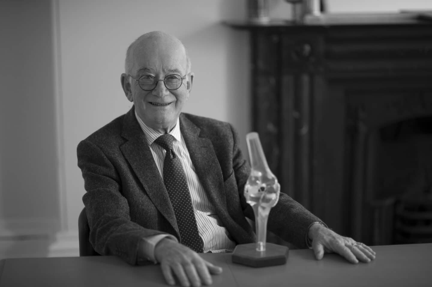 Profesor John O'Connor seated at a table wearing a suit. There is a model of a human knee (skeleton) on the table)