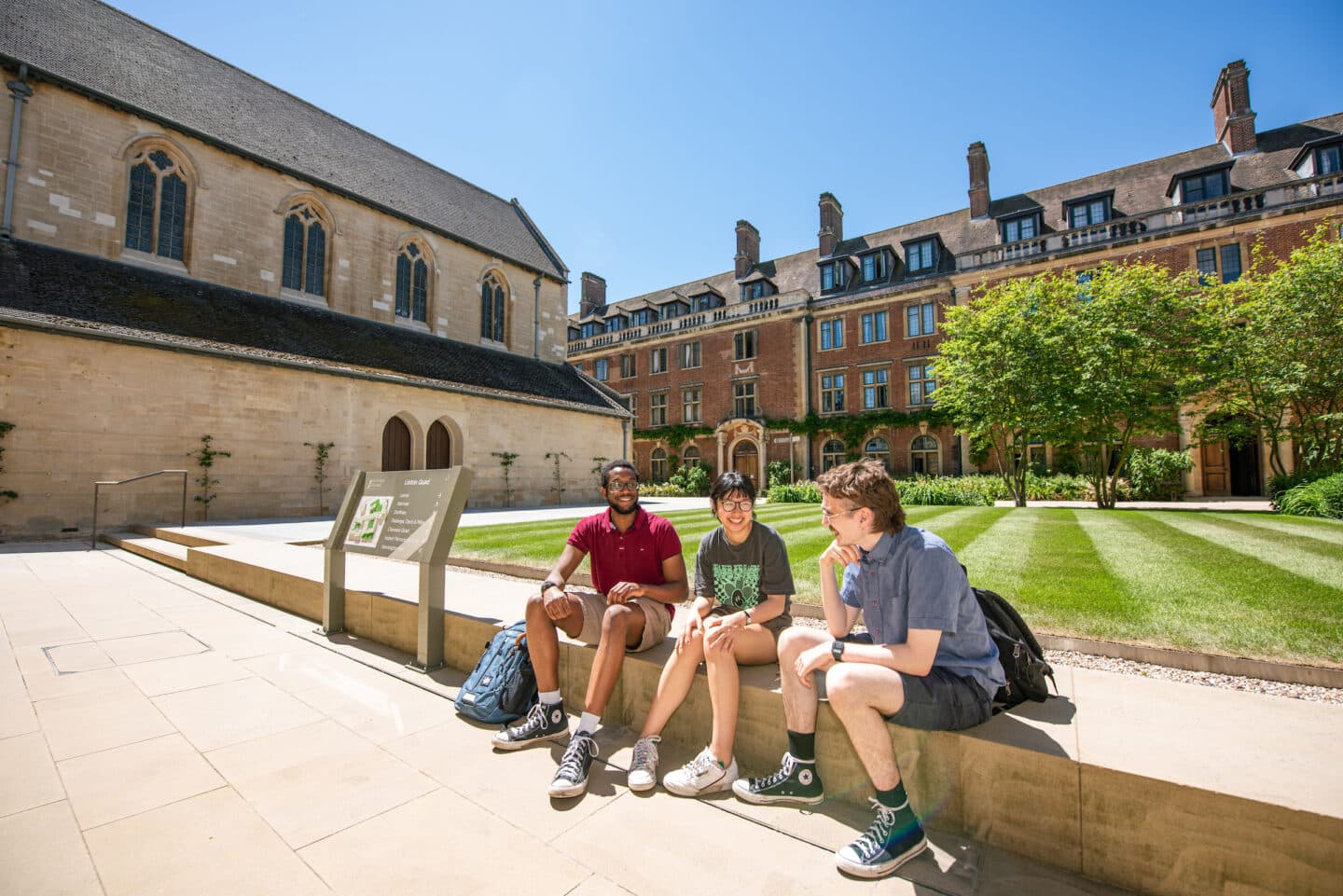 Three students seated on a concrete step with a large green lawn and old buildings behind them